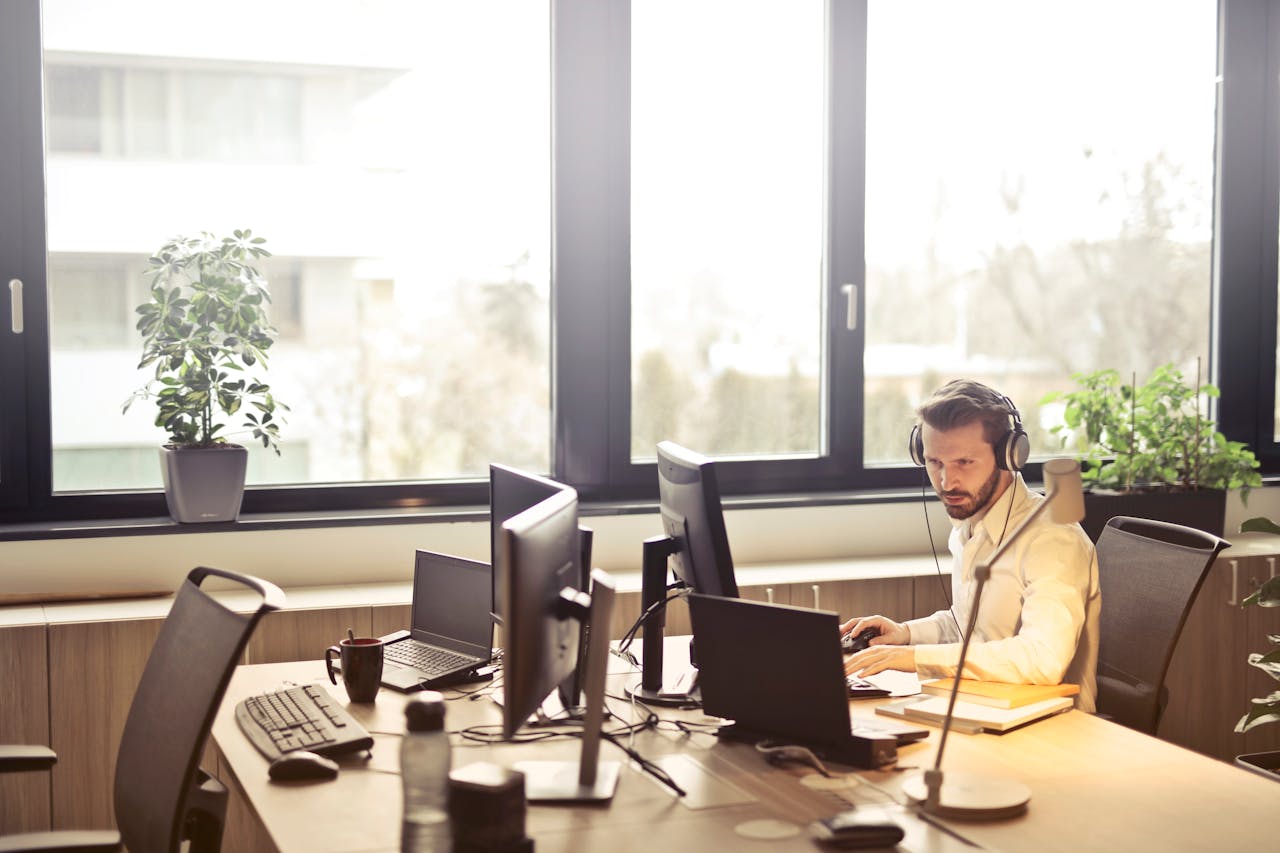 About A businessman sits at a desk using multiple computers and a headset in a well-lit modern office.